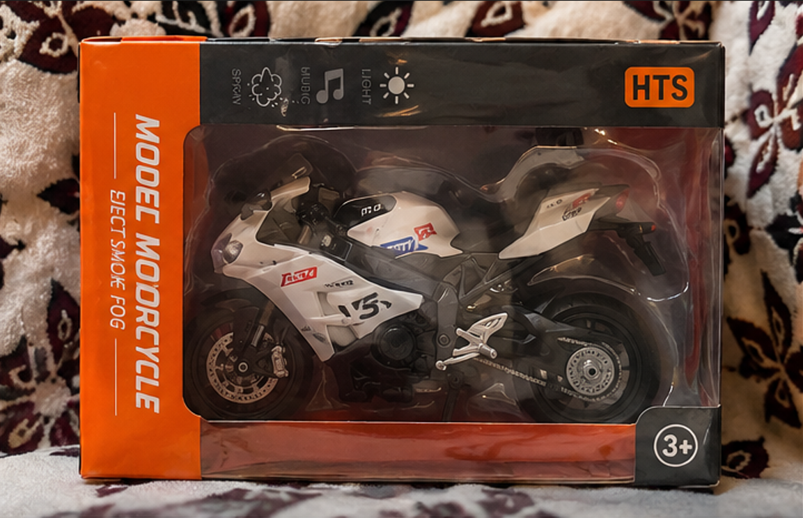 Model motorcycle toy in packaging with HTS branding on a patterned fabric background